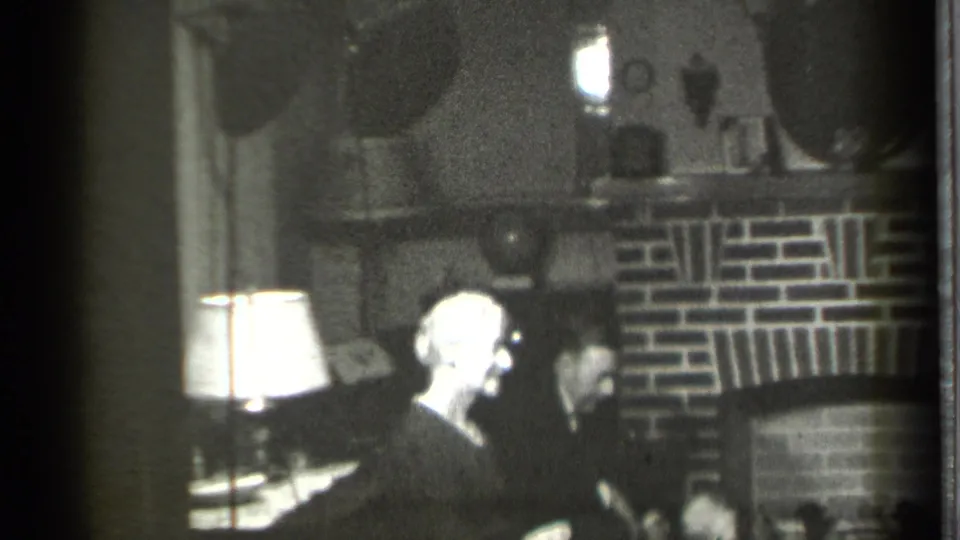 In this Danielson, Connecticut 1939 frame, a light-haired adult faces another seated person near a brick fireplace and table lamp.