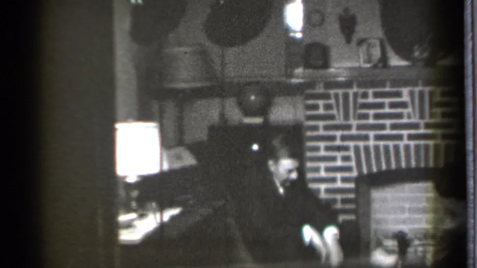 The Danielson living-room view holds on the man seated near the brick fireplace, with mantel objects and a bright lamp visible.