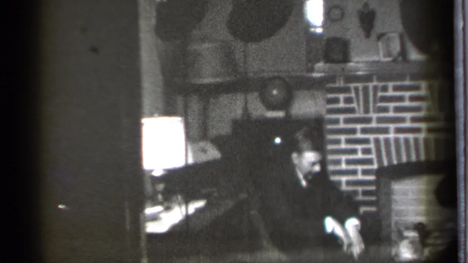 A 1939 home-movie frame shows the man seated low in the frame, facing downward in a dim Connecticut living room.