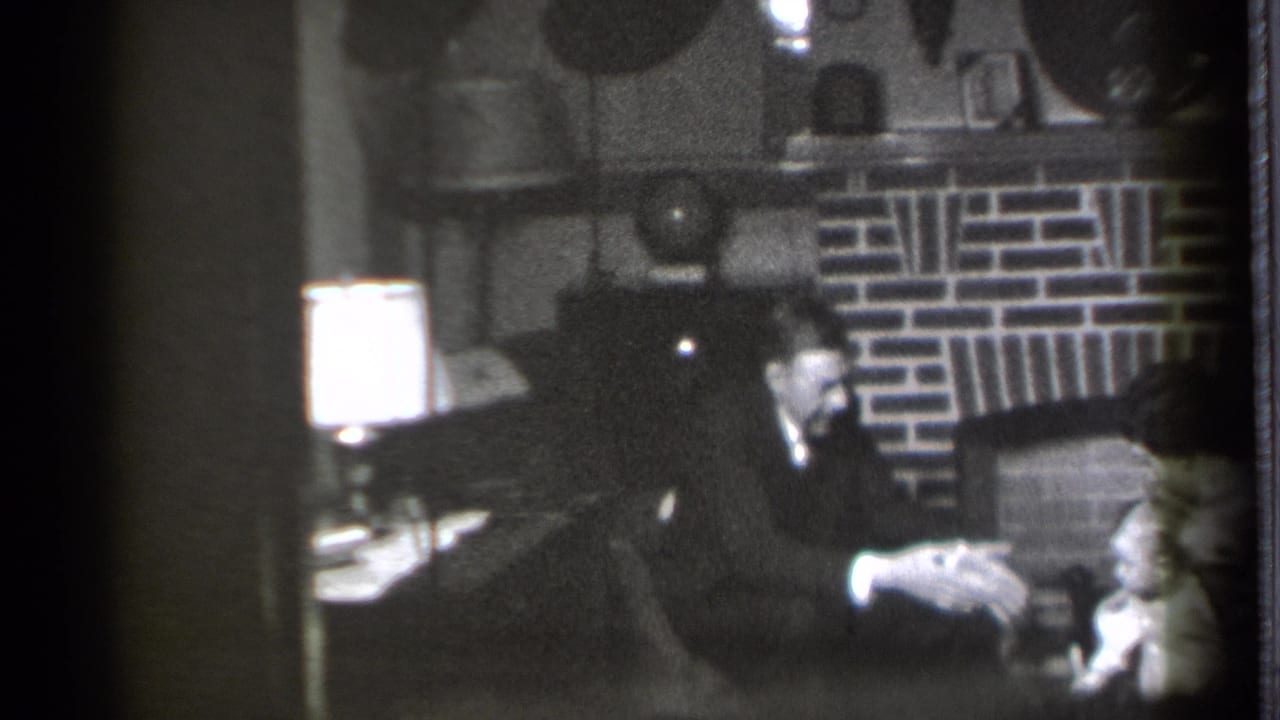 Man in a dark suit sits in a chair beside a lamp and brick fireplace, with a toddler partly visible.