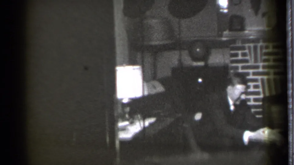 The 1939 home-movie frame shows a suited man in a den chair, a lamp at left, a brick fireplace behind him, and a child partly in view.