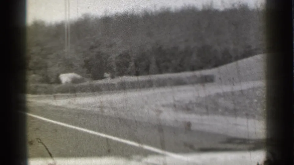 Black-and-white 8mm frame from the Danielson, Connecticut clip showing a roadside, grassy embankment, and wooded hill.