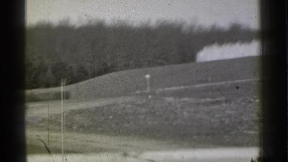 Black-and-white home-movie frame showing a quiet Danielson roadside, grassy rise, and dense tree line in soft focus.