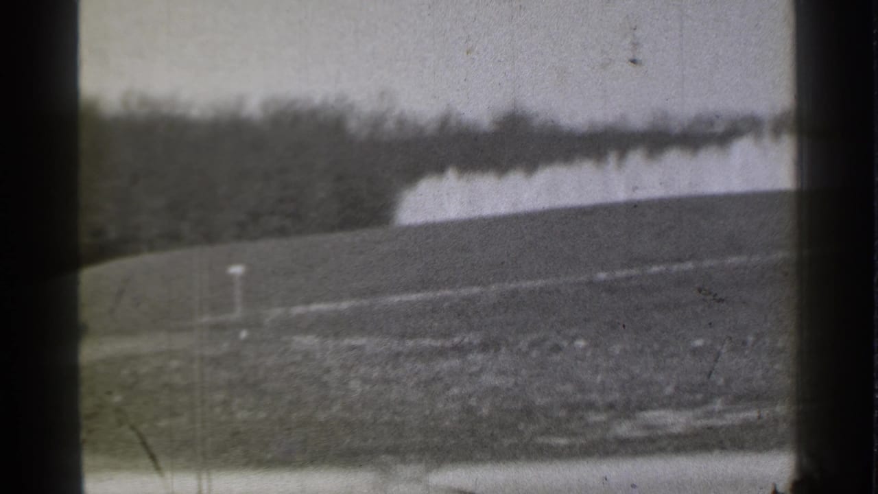 Golf course slope with a white marker post, pale water feature, and dark tree line in the distance.