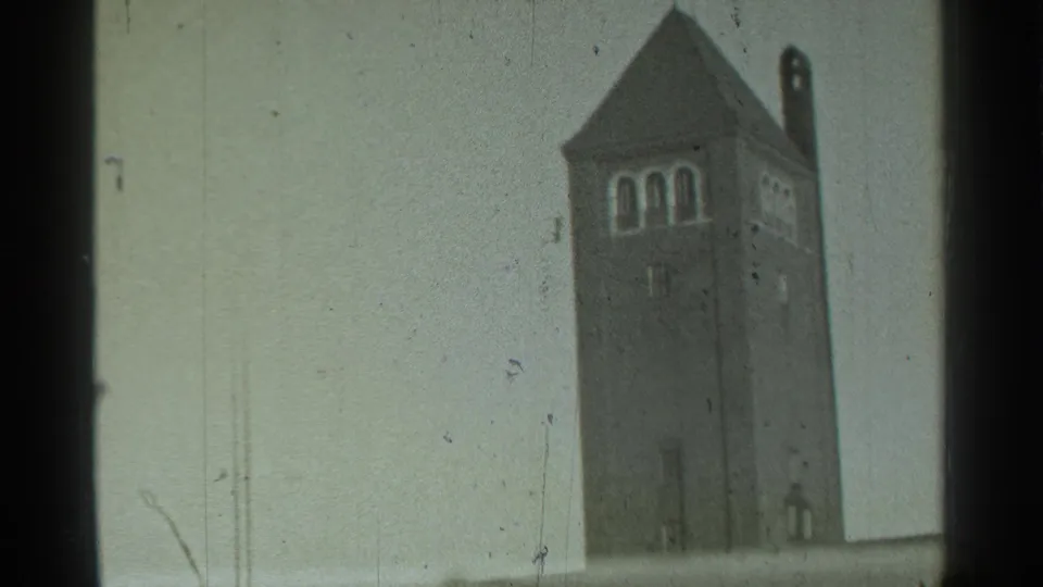 1939 Danielson, Connecticut home-movie frame showing the brick tower centered, with arched openings, pointed roof, pale sky, and lower edge blur.