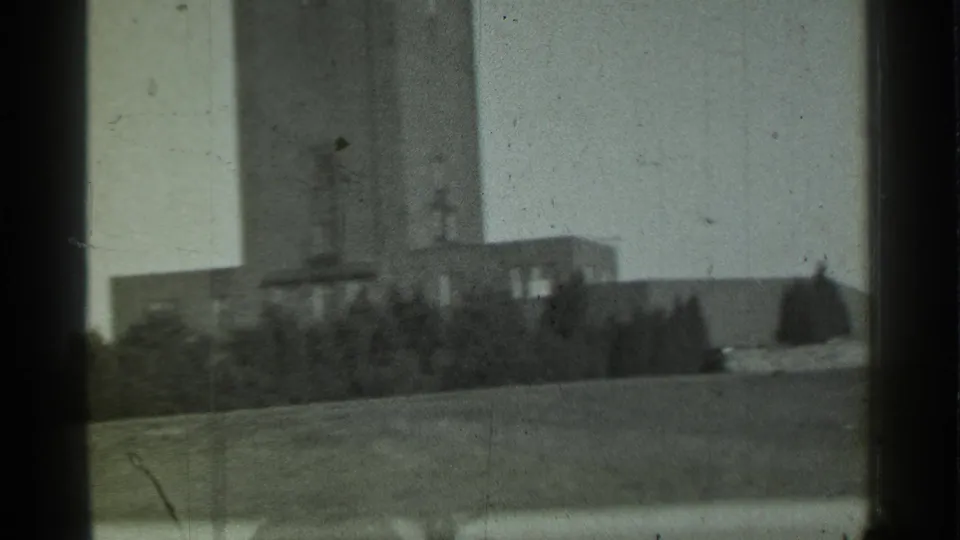 1939 Danielson, Connecticut frame showing the lower tower, adjoining low buildings, tree line, open grass, and pale sky from a moving viewpoint.