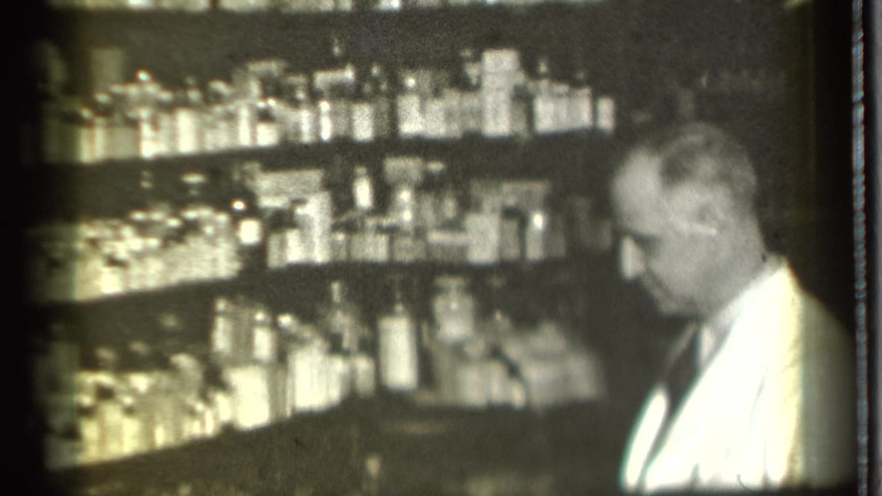 White-coated man standing beside pharmacy shelves filled with bottles and boxes.