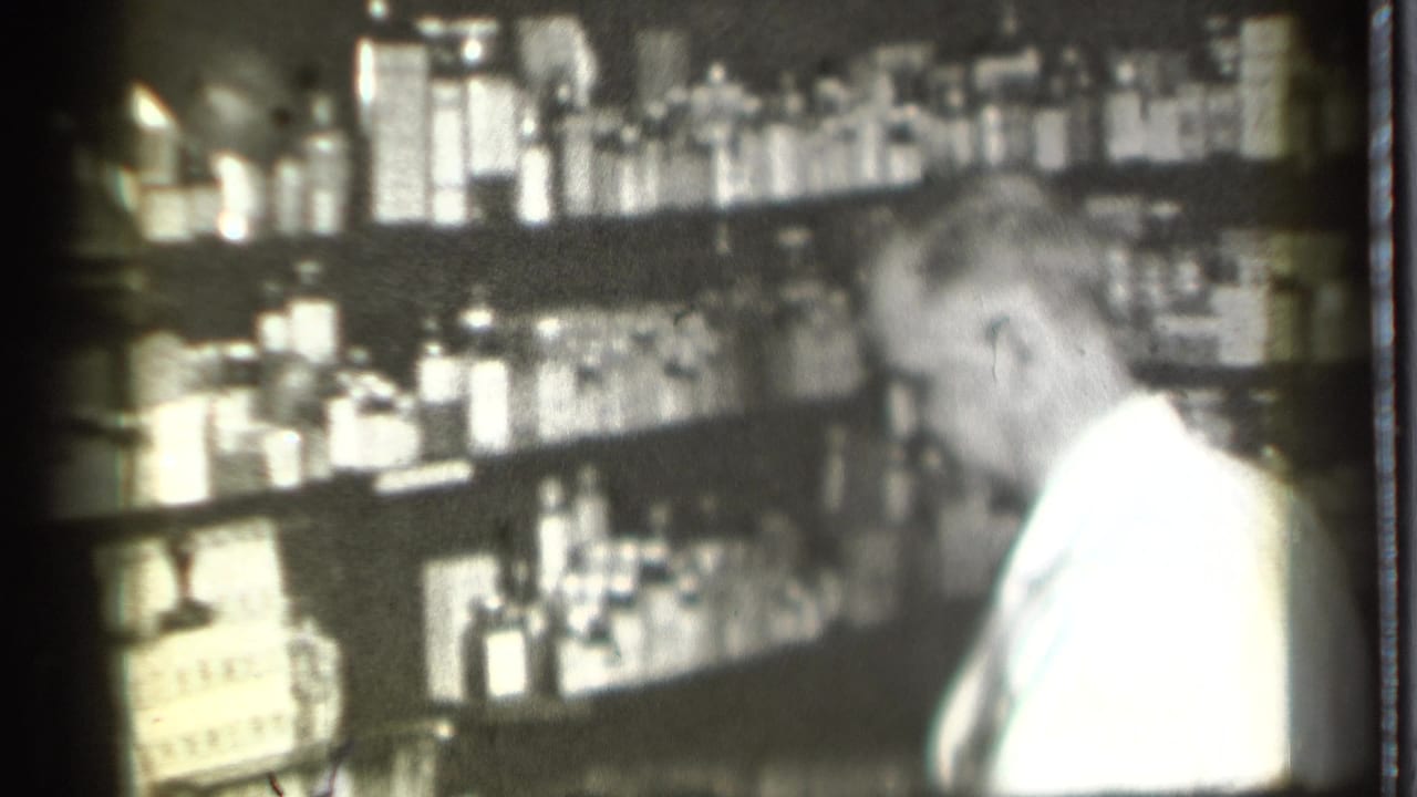 The white-coated worker turns toward shelves of bottles and packages in the Danielson pharmacy interior, filmed in soft black-and-white 8mm.