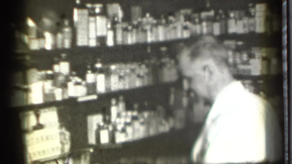 The white-coated worker turns toward shelves of bottles and packages in the Danielson pharmacy interior, filmed in soft black-and-white 8mm.