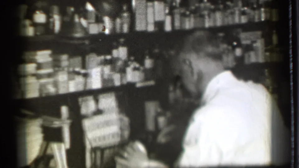 Inside the 1939 pharmacy, the man bends near the lower shelves or counter area while handling supplies among rows of bottles and boxes.