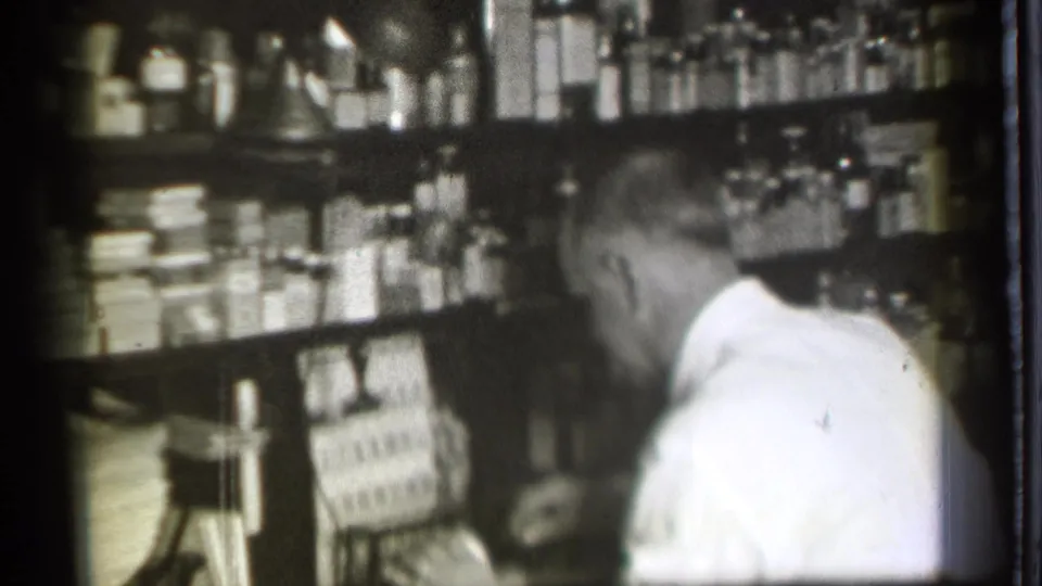 In a 1939 Danielson, Connecticut pharmacy interior, a man in a white shirt works near shelves crowded with bottles and boxed goods.