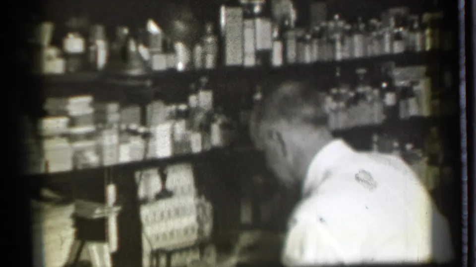 A man in a white shirt is seen from behind in the Danielson pharmacy, with stocked shelves and a counter visible around him.