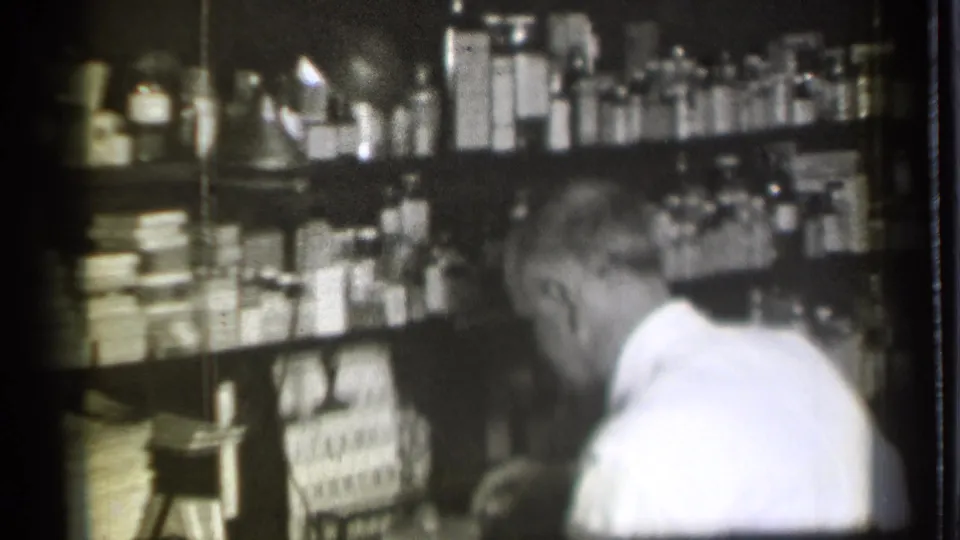 The pharmacy worker appears to bend or turn at the counter while shelves of bottles and boxes fill the background.