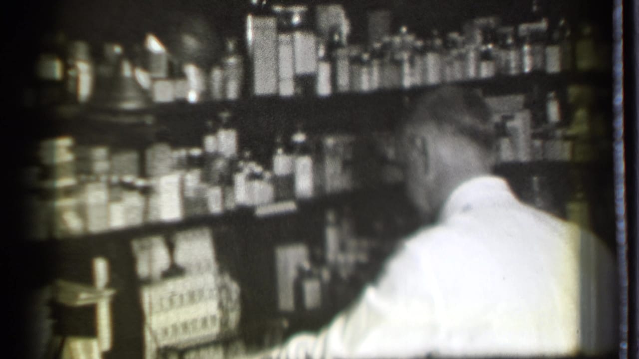 Man in a white coat working in front of crowded pharmacy shelves.