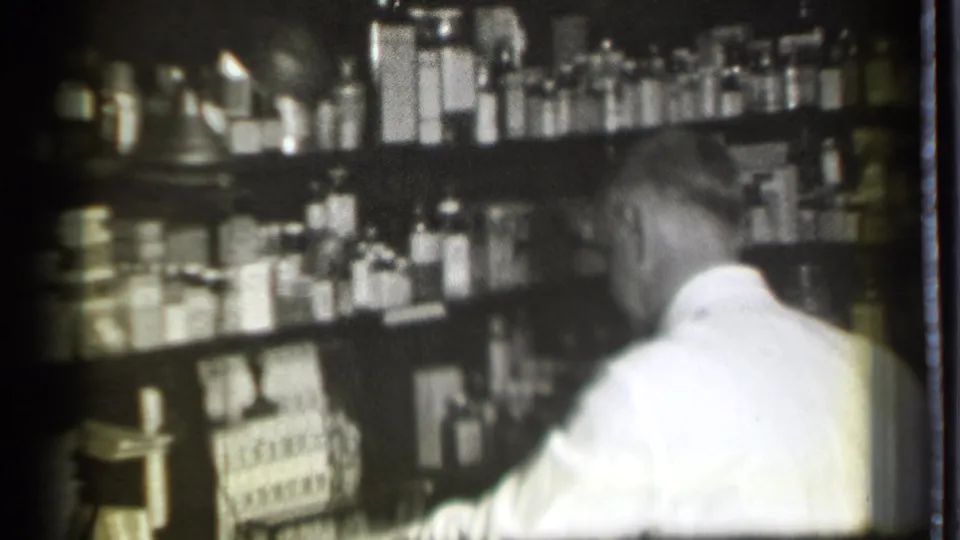 In this Danielson, Connecticut 1939 home-movie frame, a man in a white coat leans near shelves filled with bottles and boxes.