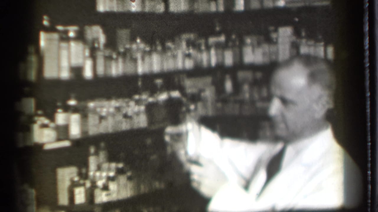 Older man in a white coat holds a glass near shelves filled with bottles.
