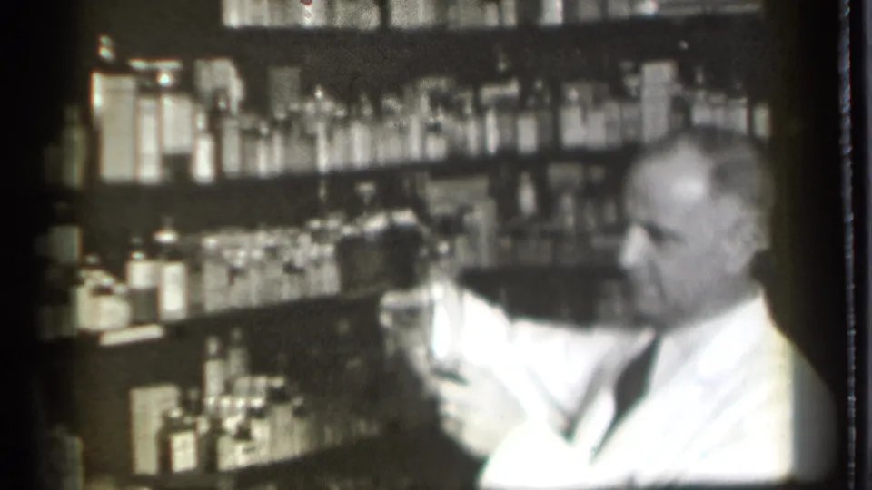 In a 1939 Danielson, Connecticut pharmacy interior, an older man in a white coat handles a glass in front of bottle-lined shelves.