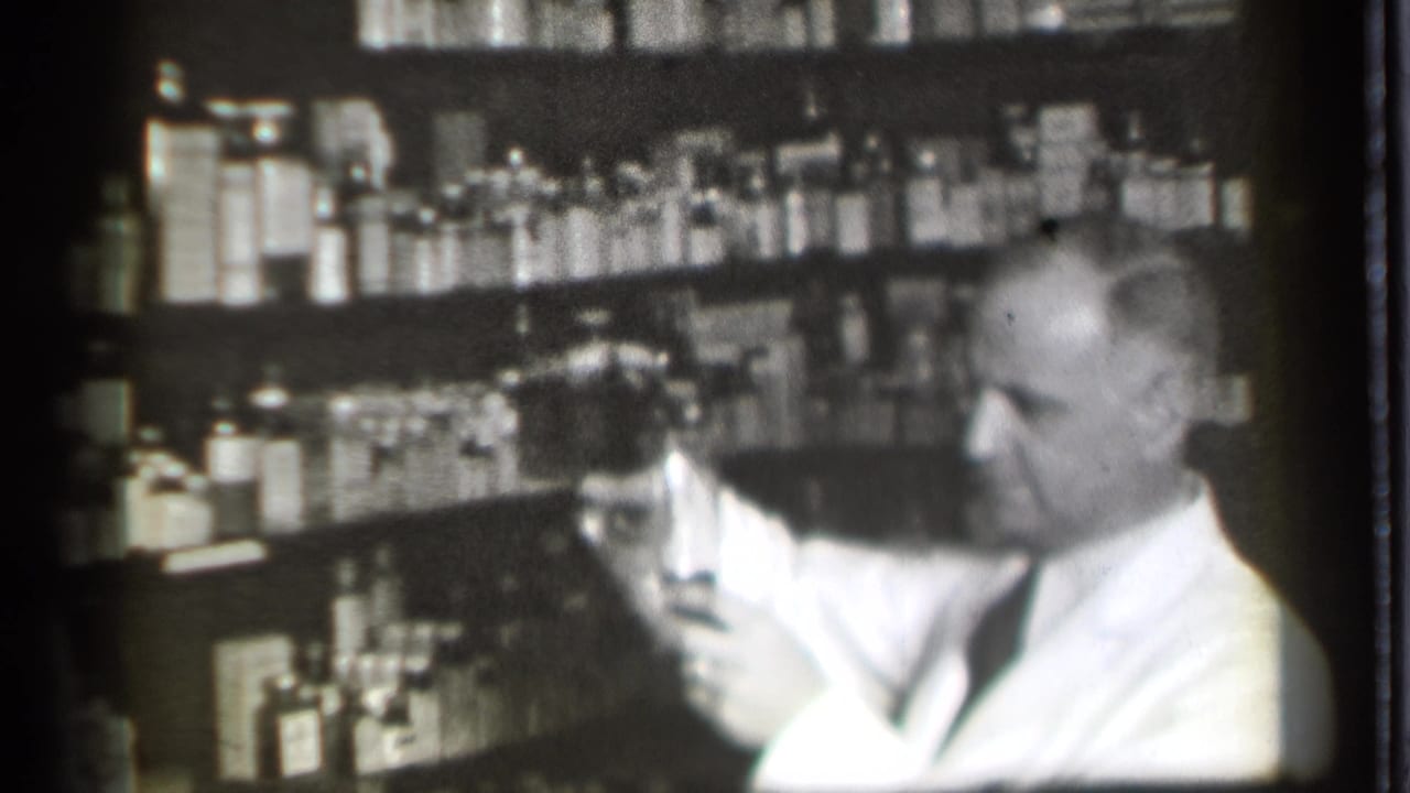 Man in a white coat looks toward a glass with pharmacy shelves behind him.