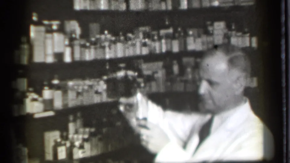 The pharmacist figure looks toward a glass while standing before crowded shelves in the Danielson pharmacy setting.