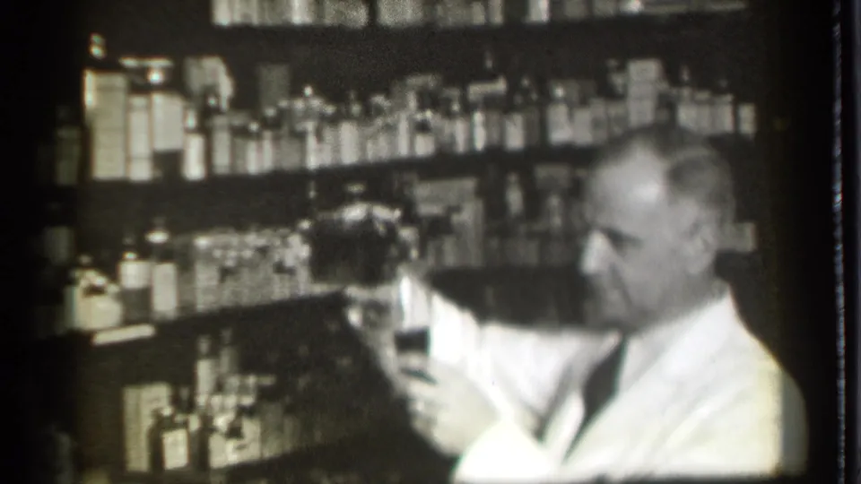 A white-coated man in the 1939 pharmacy scene lowers or steadies a glass before rows of bottles and jars.