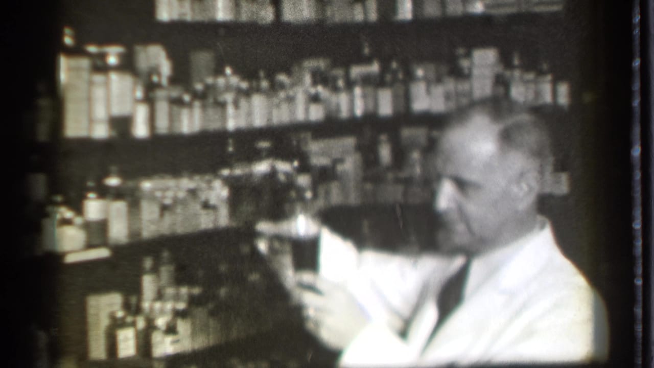 Man in a white coat measures liquid near shelves of bottles.