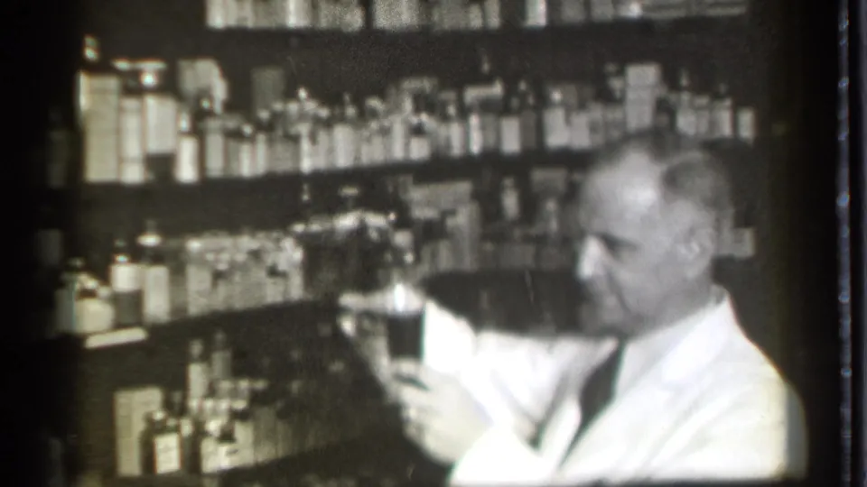 In 1939 Danielson, Connecticut footage, a man in a white coat holds glassware before shelves lined with medicine bottles.