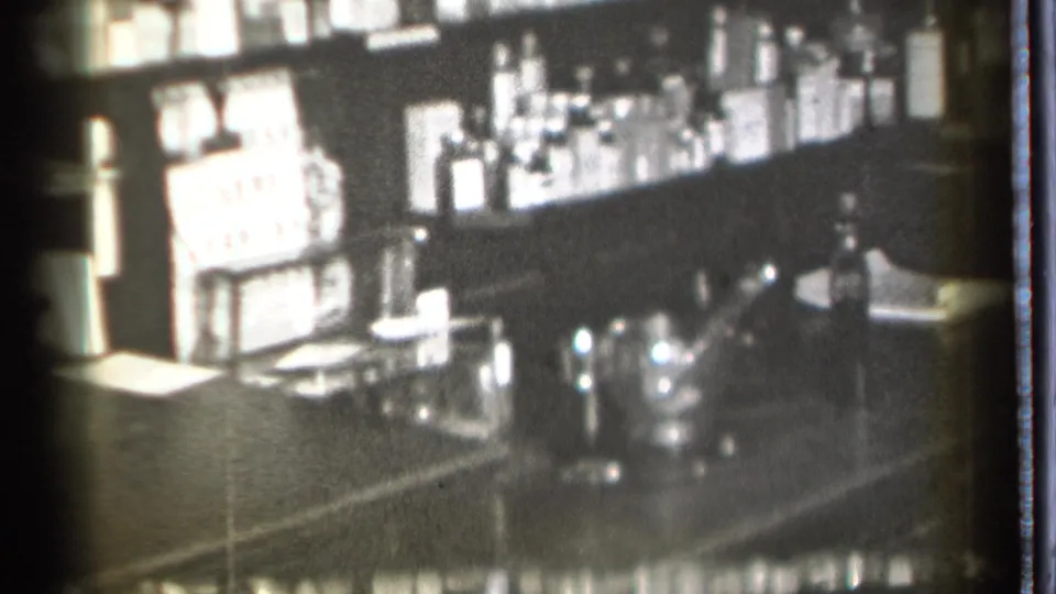 Soft black-and-white 8mm frame of a Danielson bar interior, with bottles on shelves and glassware clustered near the counter.