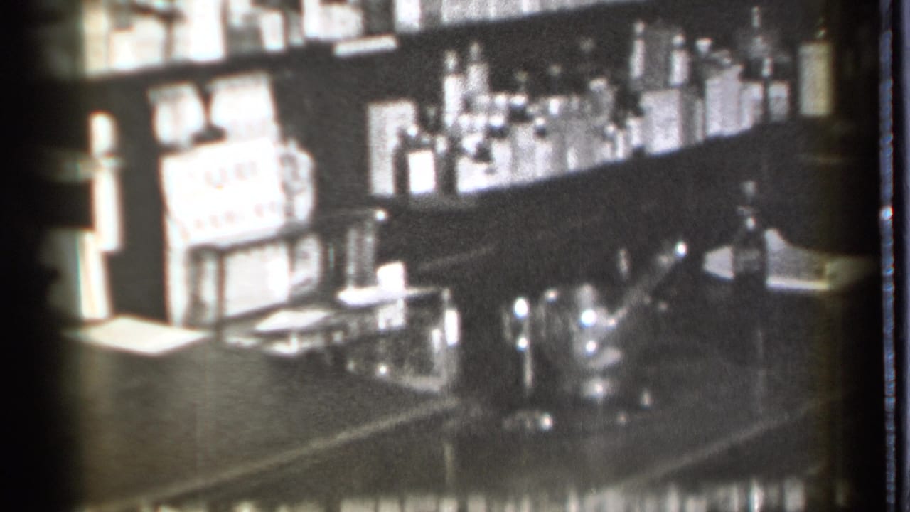Pharmacy counter and shelves filled with bottles in soft black-and-white footage.