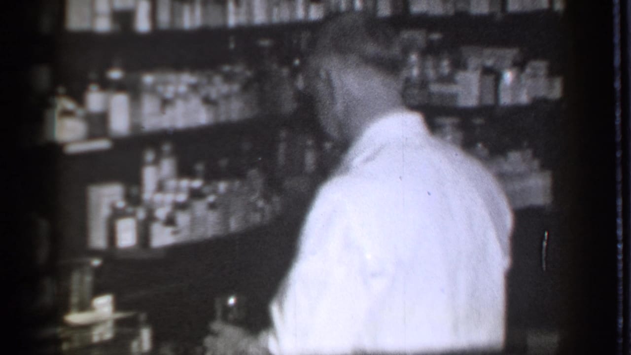 Man in a white lab coat stands behind a pharmacy counter facing shelves of bottles.