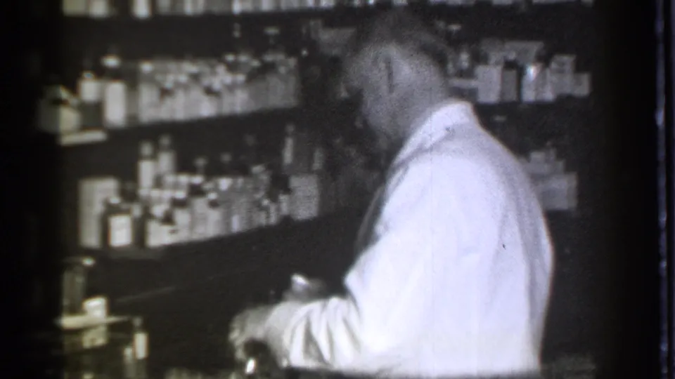 In the 1939 Danielson pharmacy, the pharmacist looks down while handling items near the counter, with rows of bottles behind him.