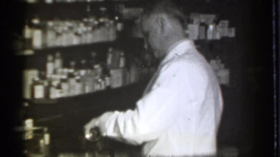 A white-coated pharmacist works at the counter inside a 1939 Danielson drugstore, surrounded by shelves of glass bottles.