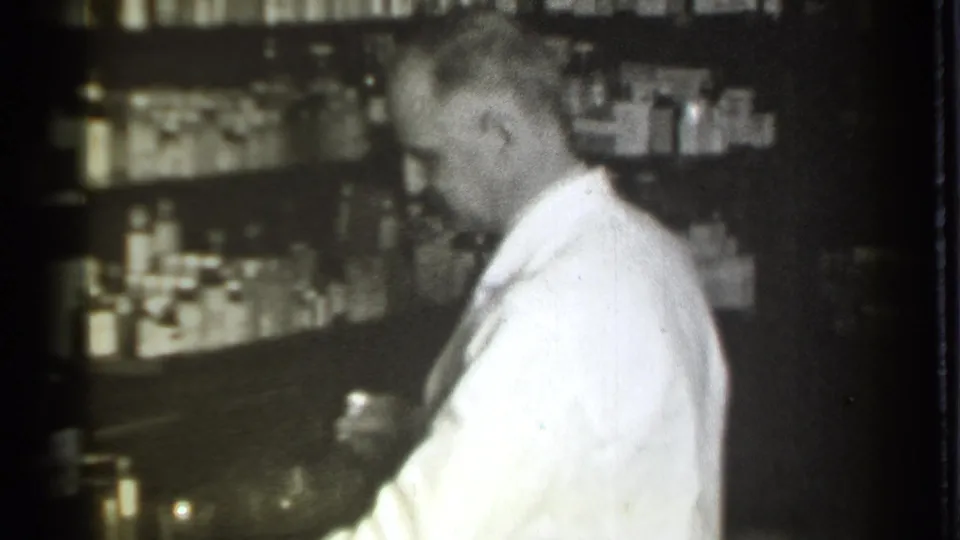 The pharmacist looks down at a small container in the 1939 Danielson pharmacy, with rows of bottles visible on shelves behind him.