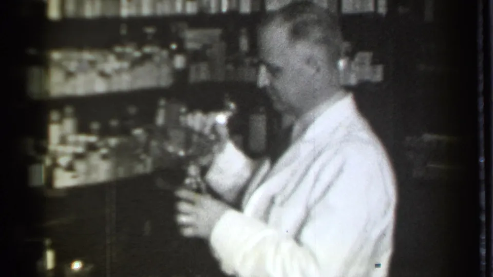 The pharmacist raises a glass bottle or jar in the Danielson, Connecticut pharmacy, with stocked shelves filling the background.