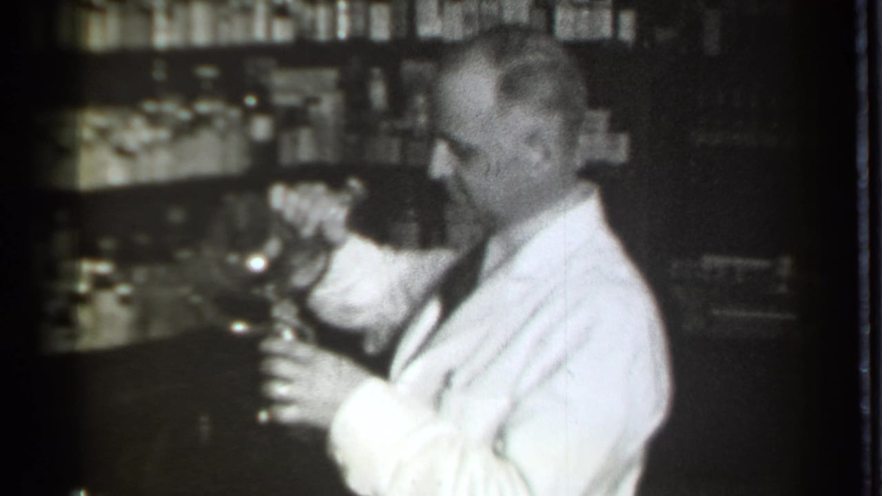 Man in a white coat holds a glass vessel behind a drugstore counter.