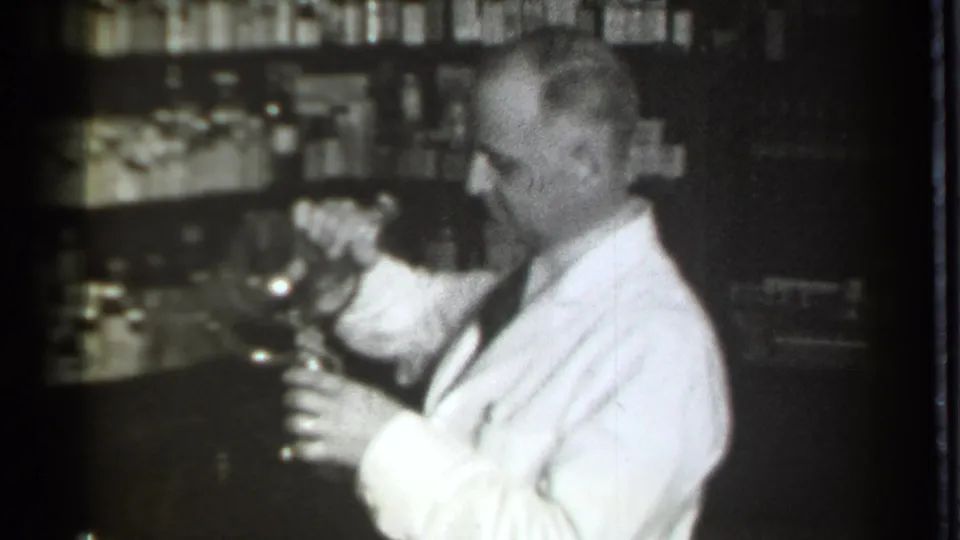 In Danielson, Connecticut, 1939 footage, a pharmacist in a white coat handles a glass vessel with shelves of bottles behind him.