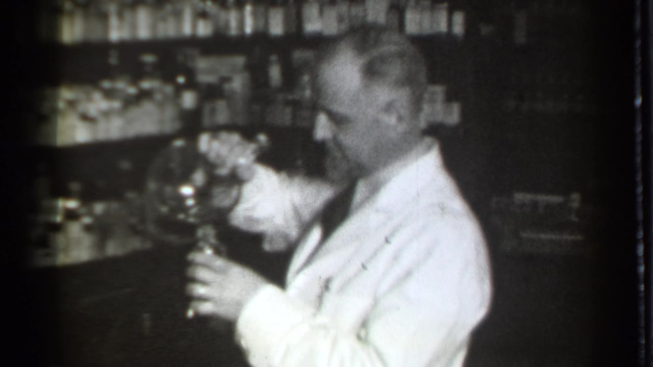 White-coated man looks down while handling glassware near shelves of bottles.