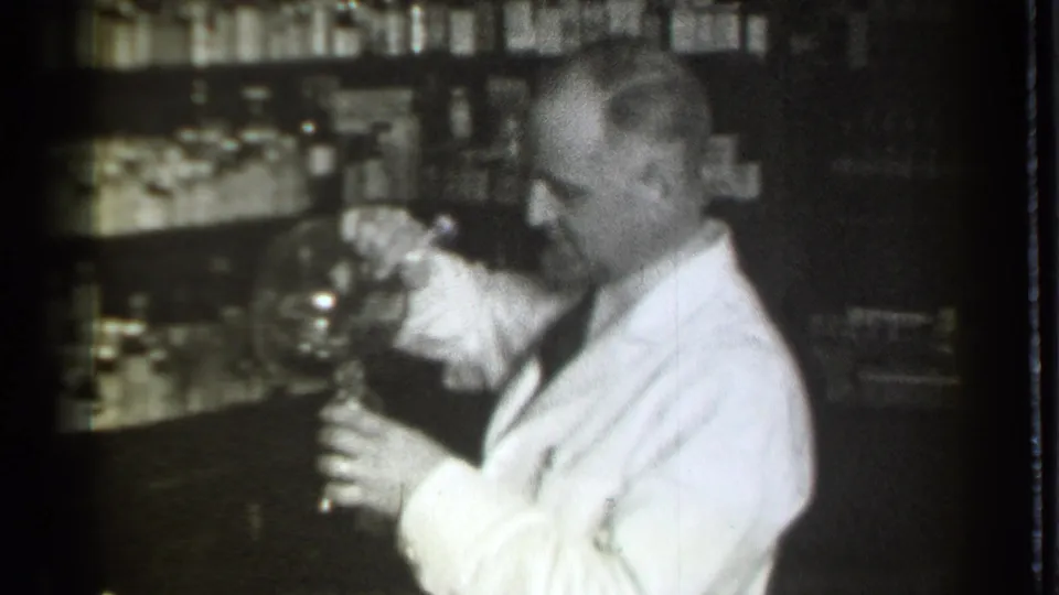A white-coated pharmacist in the Danielson, Connecticut drugstore continues working with glassware against a backdrop of medicine bottles.
