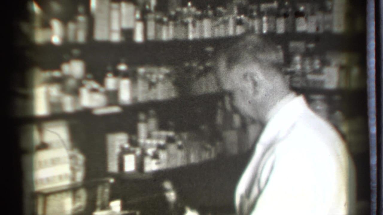 Man in a white lab coat stands beside shelves of pharmacy bottles.