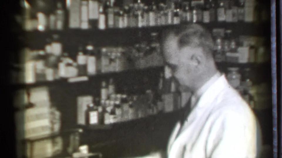 A pharmacist in a white coat is seen in profile inside the 1939 Danielson pharmacy, surrounded by medicine bottles.
