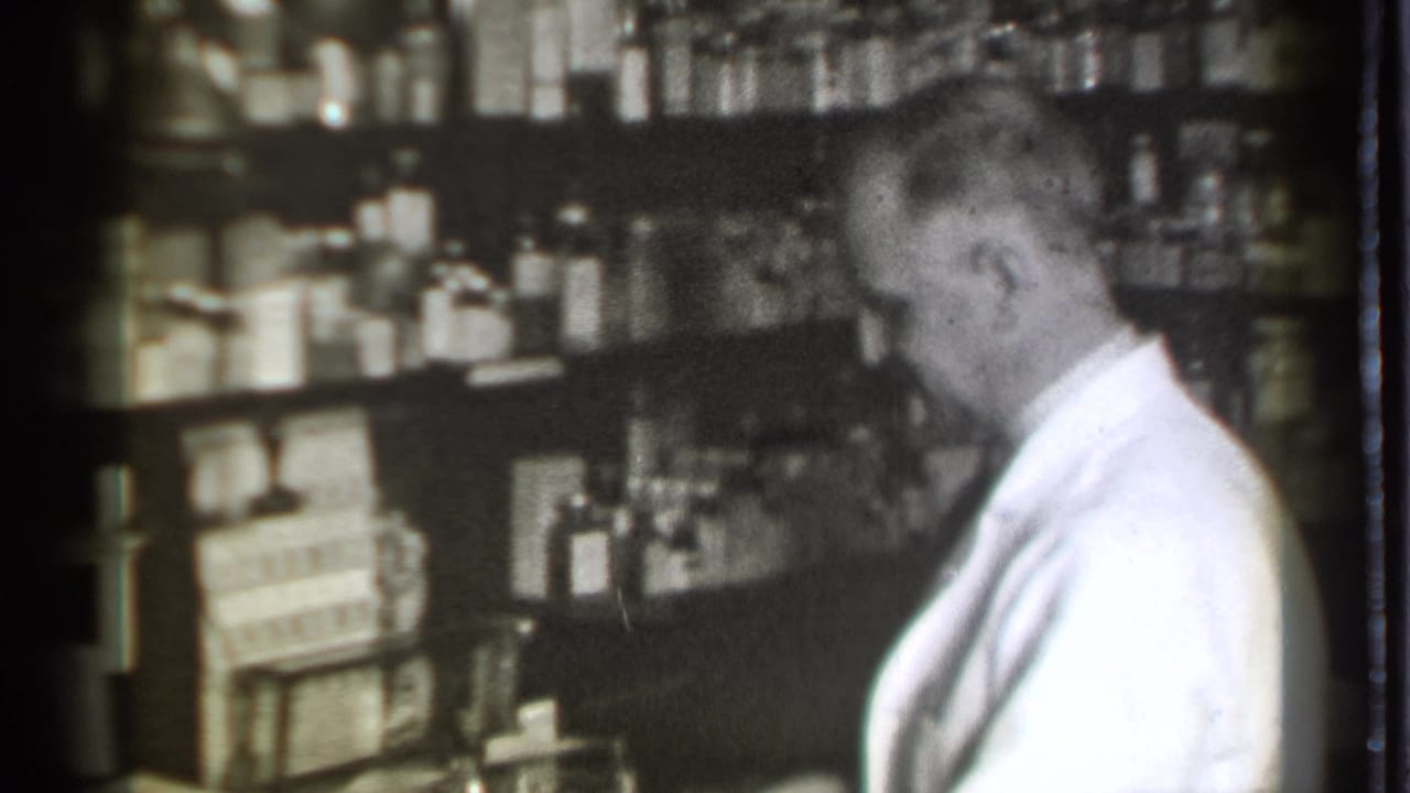 Pharmacist in a white coat stands beside shelves filled with bottles and boxes.