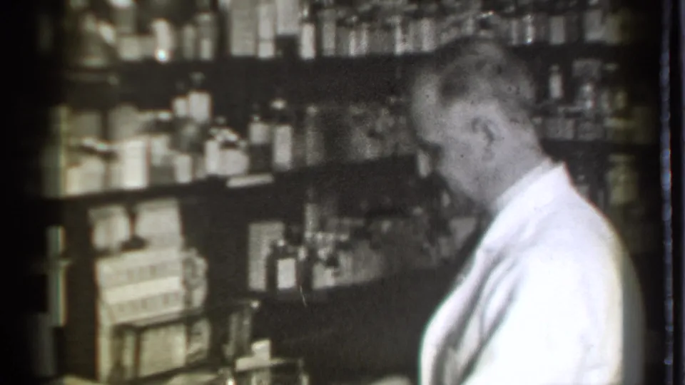 In Danielson, Connecticut, 1939, a pharmacist in a white coat works near packed drugstore shelves of bottles and boxed goods.