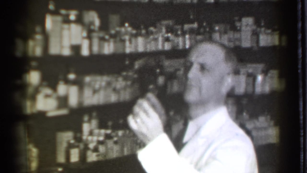 Pharmacist holds a small bottle or container in front of shelves of supplies.