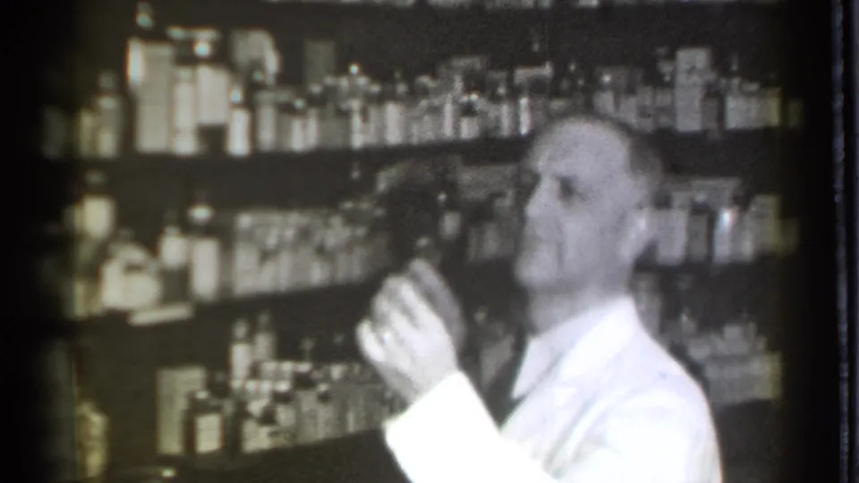 In the 1939 Danielson drugstore footage, the pharmacist raises a small container while standing before shelves of medicine bottles.