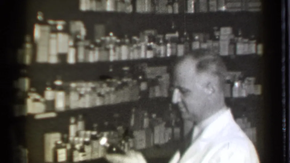 The Danielson pharmacist appears to handle a small item at the counter, with rows of glass bottles and supplies behind him.