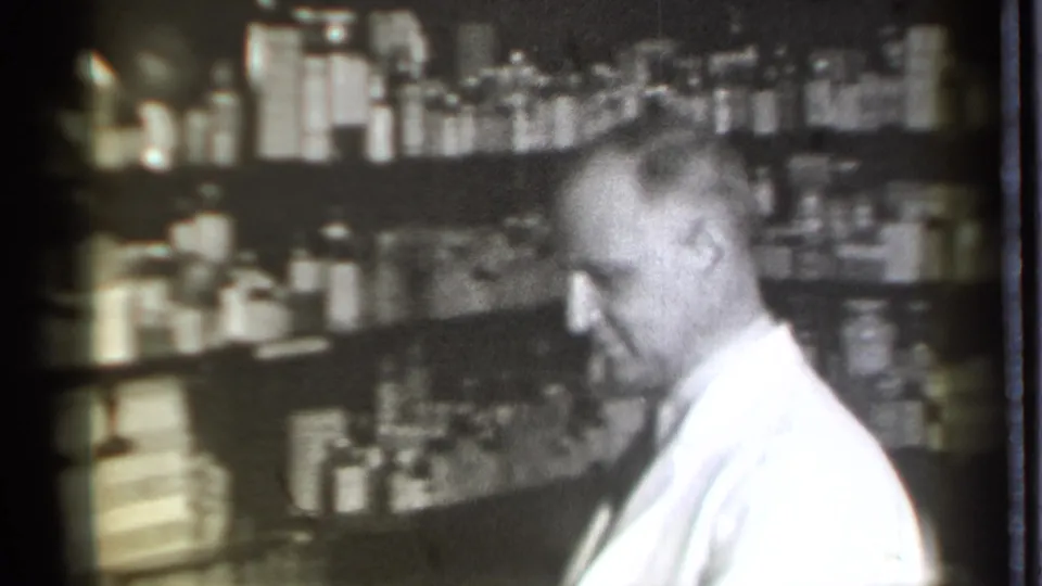 A pharmacist in a white coat looks downward in the 1939 Danielson drugstore, framed by shelves of medicine bottles and packaged goods.
