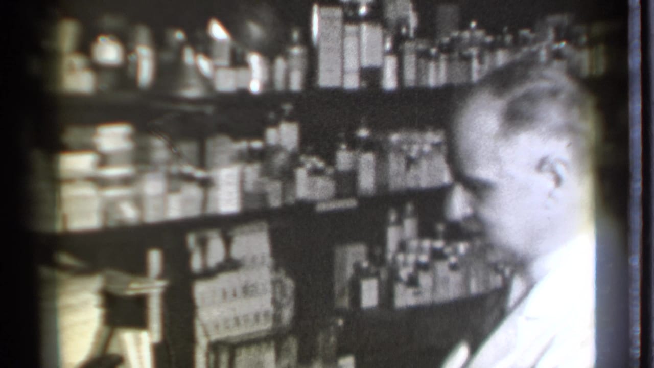Pharmacist in a white coat beside shelves filled with bottles and medicine boxes.