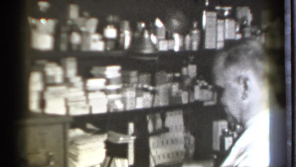 A pharmacist in a white coat leans near the counter in a Danielson, Connecticut pharmacy, surrounded by shelves of medicines and supplies.