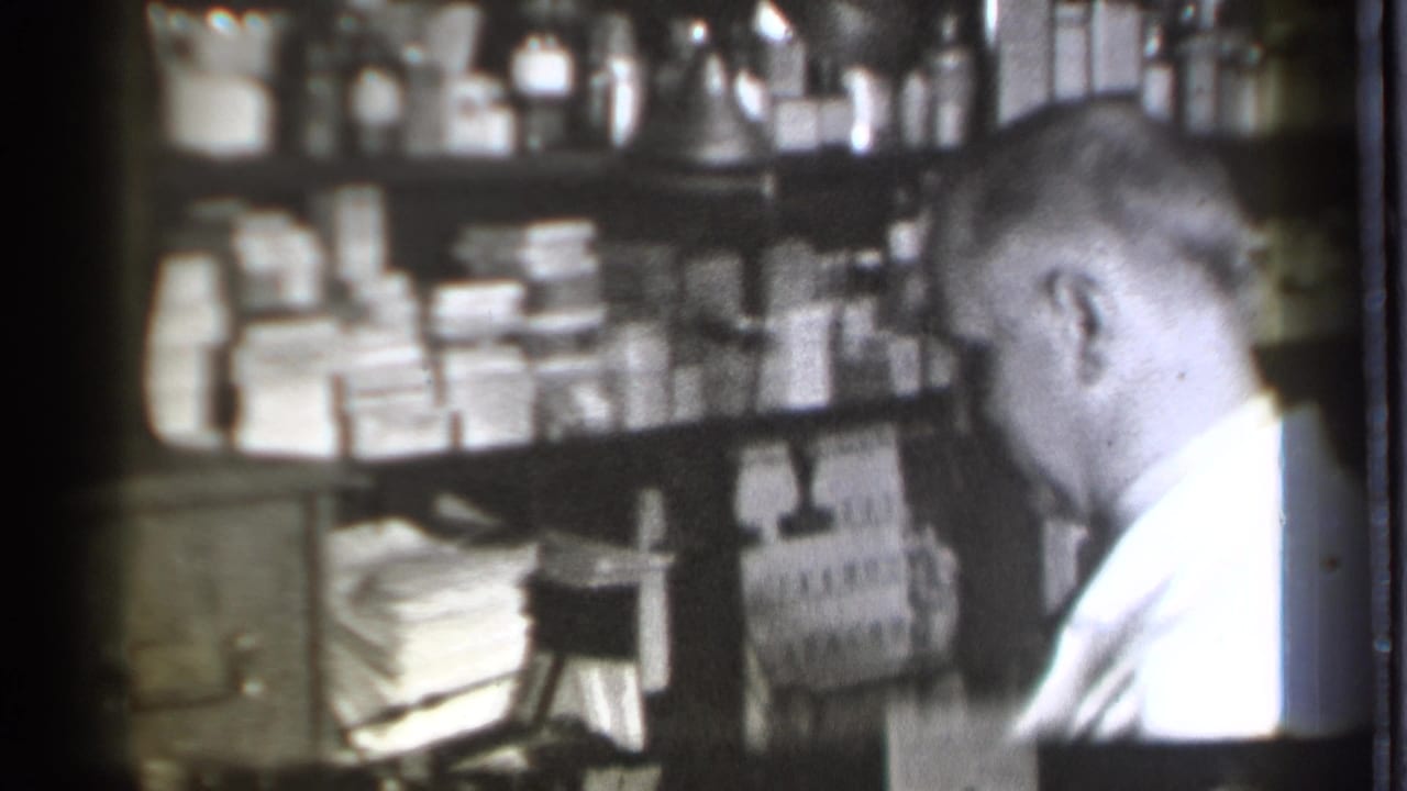 Man in white coat looks toward stocked pharmacy shelves and boxed medicines.