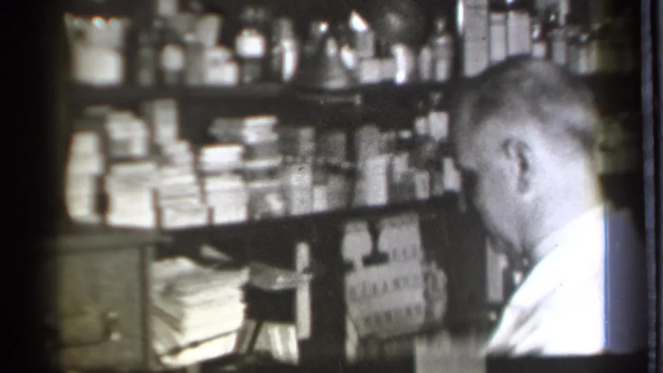 The pharmacist remains beside the Danielson drugstore shelves, with boxed medicines and jars visible across the black-and-white 8mm frame.