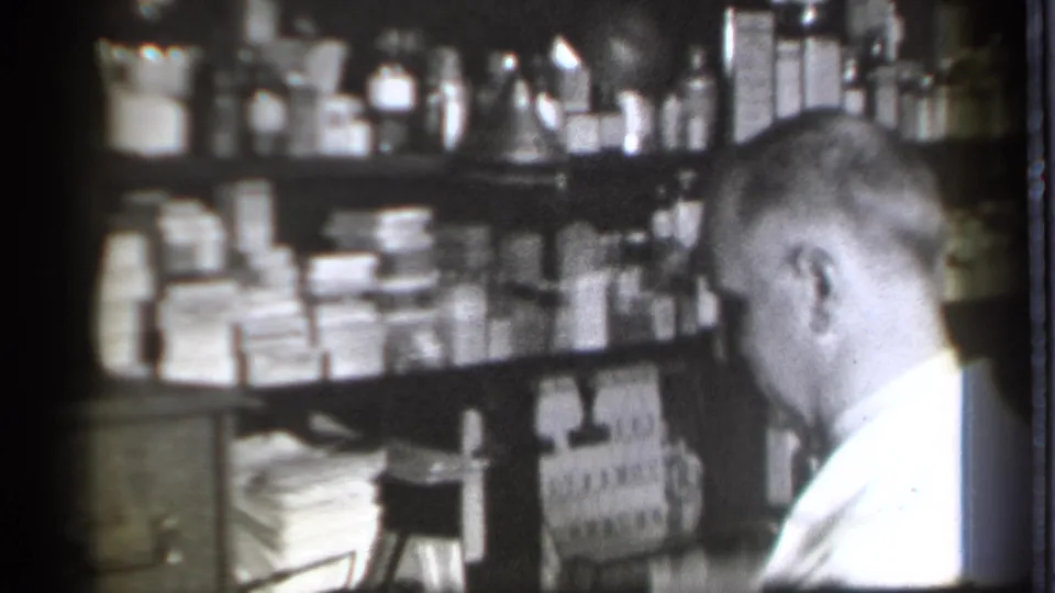 At the end of the clip, the pharmacist bends near dense shelves of medicine containers in the 1939 Danielson, Connecticut drugstore interior.